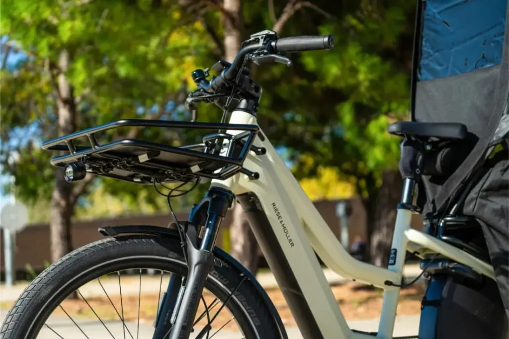 Primer plano de una bicicleta eléctrica de carga Riese & Müller Multicharger3 Mixte Vario con cesta delantera, aparcada al aire libre cerca de unos árboles.