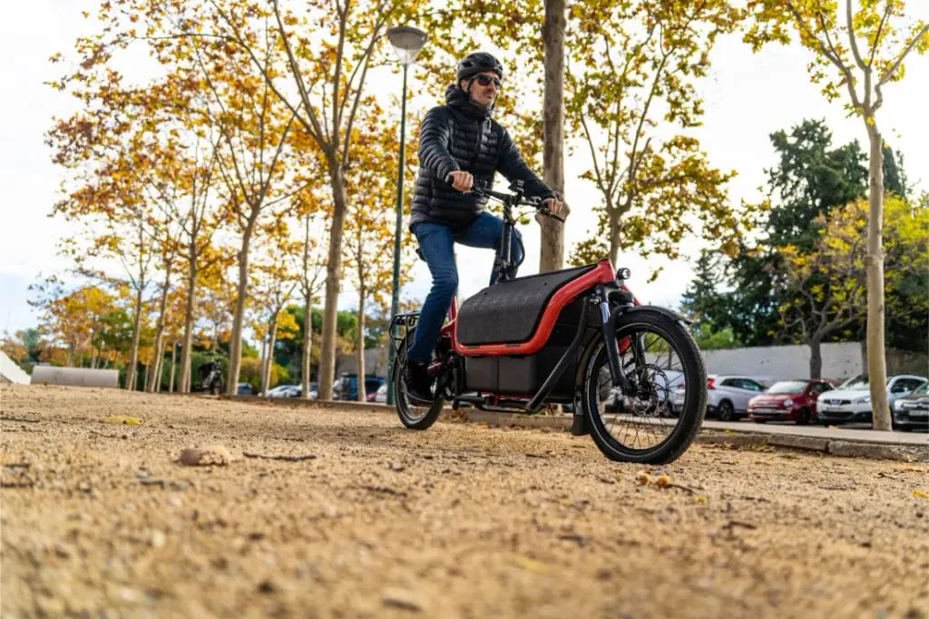 Una persona monta en una bicicleta de carga Riese Müller Carrie2 por un camino de tierra bordeado de árboles otoñales y coches aparcados al fondo, disfrutando de su cuadro compacto y su facilidad automática.
