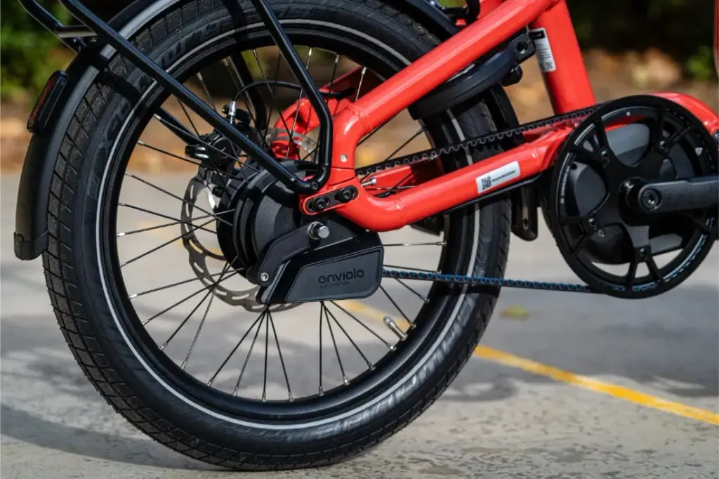 Primer plano de la rueda trasera y el mecanismo de la cadena de una bicicleta eléctrica automática Riese Müller Carrie2 roja sobre una acera, mostrando su diseño compacto.