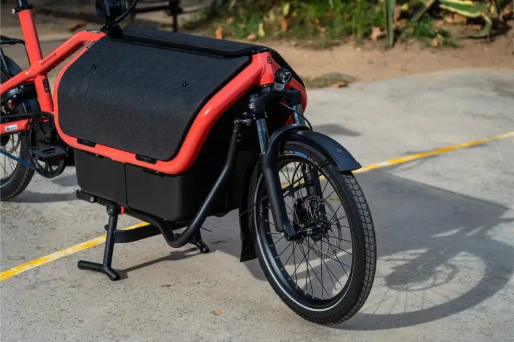 Primer plano frontal de la bicicleta eléctrica de carga compacta Riese Müller Carrie2 roja, con compartimento de almacenamiento automático negro y pata de cabra en el pavimento.