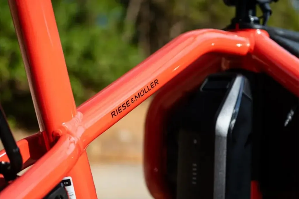 Primer plano de un cuadro de bicicleta compacto de color naranja con la inscripción "Riese & Müller", batería parcialmente visible: destaca el moderno diseño automático Carrie2.