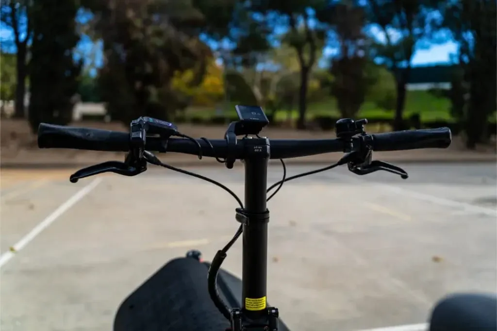 Primer plano del manillar de una bicicleta compacta Riese Müller en un aparcamiento vacío, enmarcado por árboles al fondo.