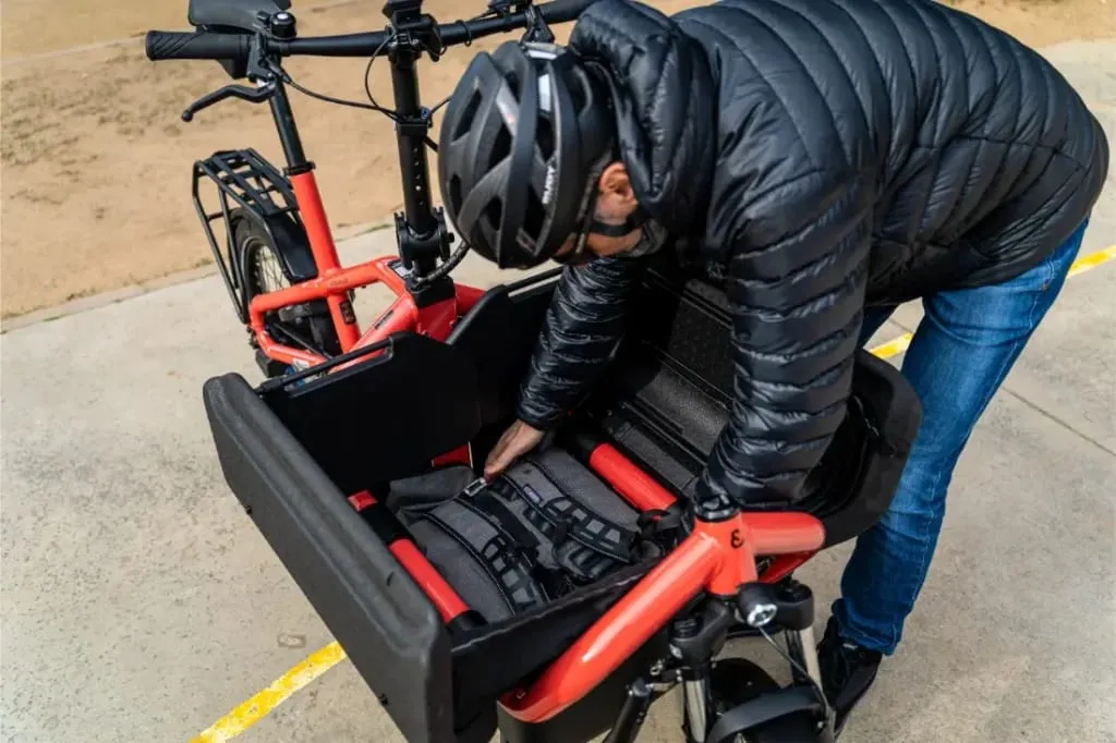 Una persona con casco y chaqueta se asegura el cinturón de seguridad en la zona de carga delantera de un Riese Müller Carrie2 eléctrico compacto rojo.