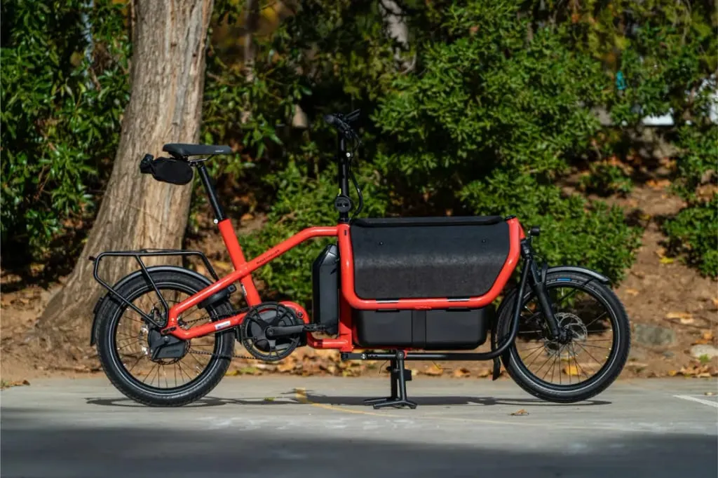 Una bicicleta de carga compacta automática Riese Müller Carrie2 de color rojo aparcada en la acera con vegetación y árboles al fondo.