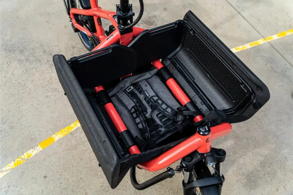 Un primer plano de la caja de carga delantera de un compacto Riese Müller Carrie2 con una mochila gris asegurada en su interior.