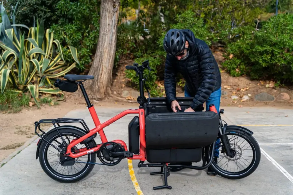 Una persona con casco ajusta el compartimento portaobjetos de una bicicleta eléctrica de carga Riese Müller Carrie2 roja, conocida por su tamaño compacto, aparcada al aire libre.