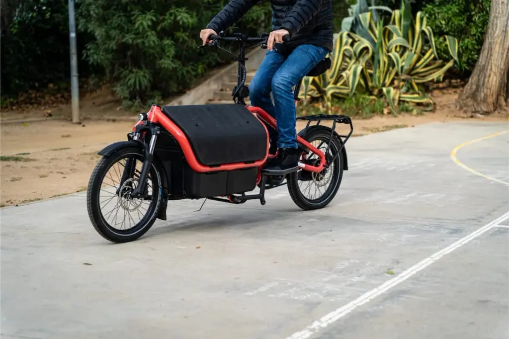 Una persona conduce una bicicleta eléctrica de carga automática Riese Müller Carrie2 roja por un sendero exterior pavimentado, rodeado de exuberante vegetación.