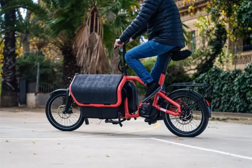 Una persona conduce una bicicleta eléctrica de carga automática Riese Müller Carrie2 roja sobre una superficie pavimentada, rodeada de árboles y vegetación, mostrando su diseño compacto para la movilidad urbana.