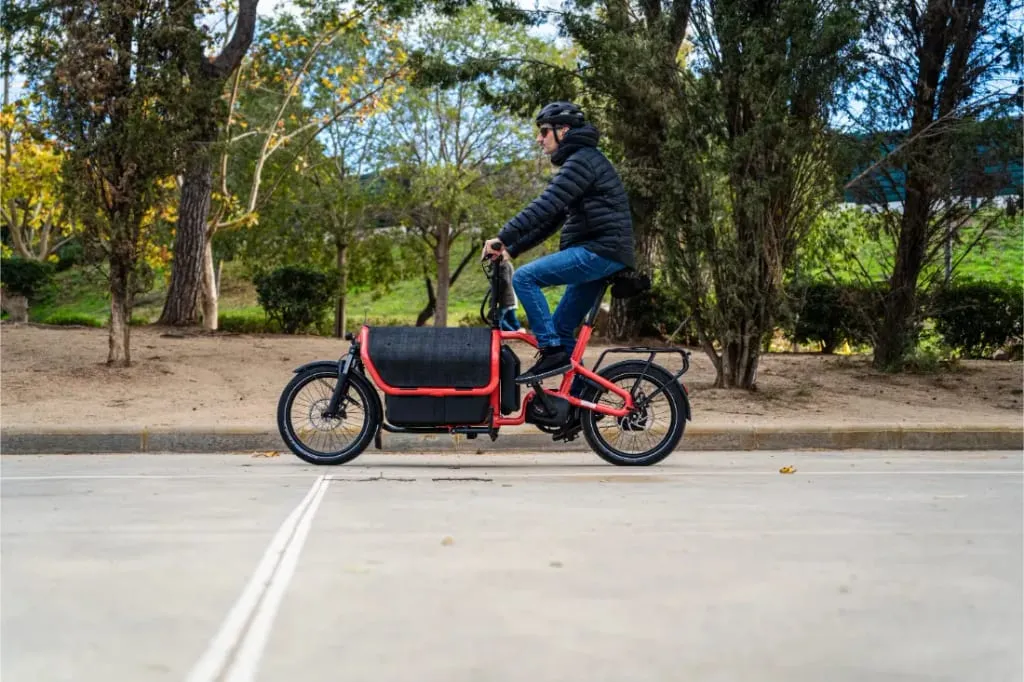 Una persona con casco conduce una bicicleta de carga compacta Riese Müller Carrie2 con cambio automático por un camino pavimentado de un parque.
