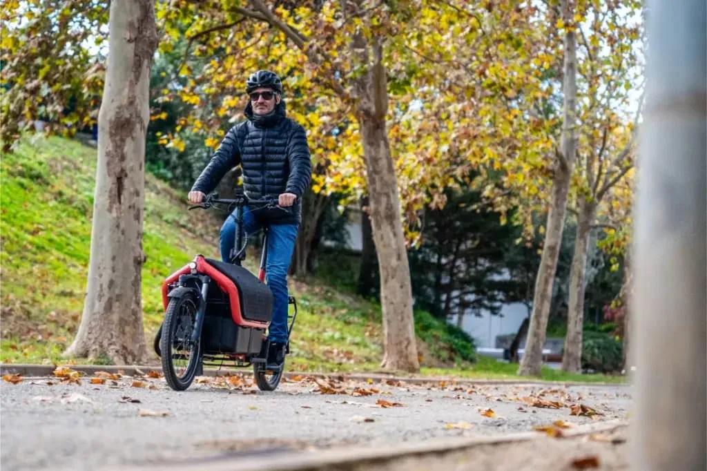 Una persona conduce una bicicleta de carga Riese Müller Carrie2 de tamaño compacto por un camino arbolado, con hojas otoñales esparcidas por el suelo.