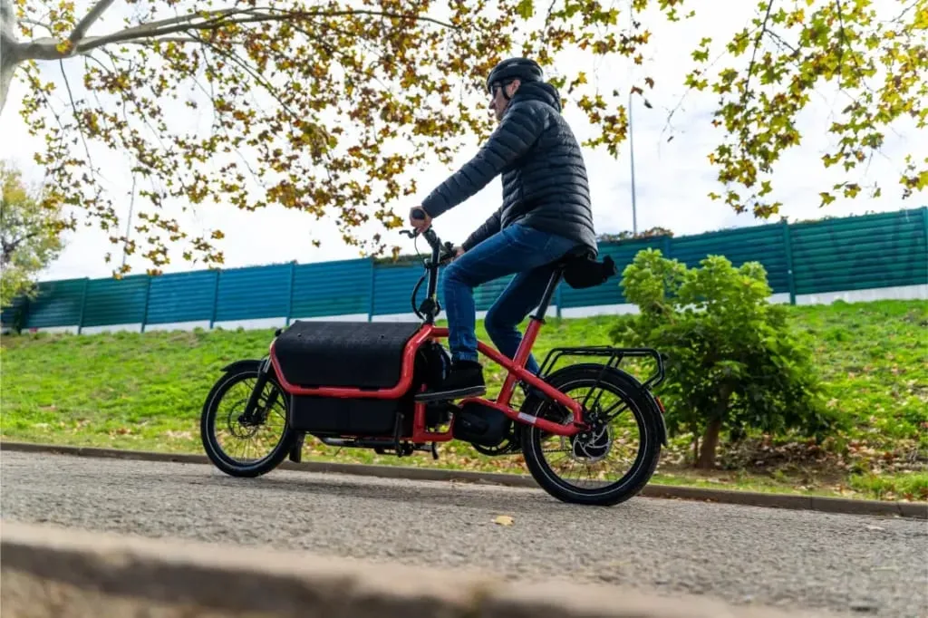 Una persona con casco y chaqueta conduce una Riese Müller Carrie2, una bicicleta de carga roja automática de tamaño compacto, por un camino asfaltado cerca de una zona de hierba con árboles.