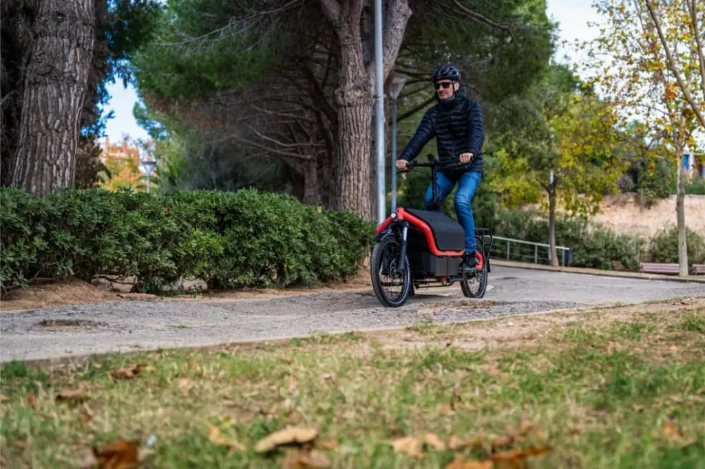 Una persona con casco conduce una bicicleta eléctrica Riese Müller roja por un camino pavimentado a través de un parque con árboles.