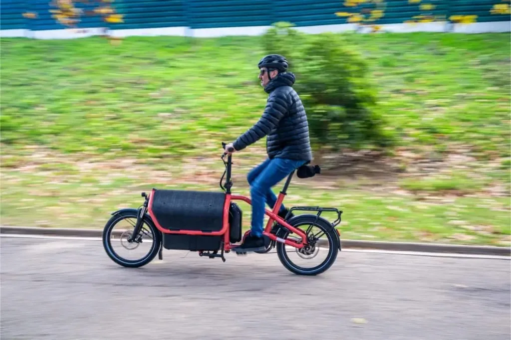 Hombre montado en una bicicleta de carga Riese Müller Carrie2 de tamaño compacto en una carretera, con casco y chaqueta, con vegetación de fondo.