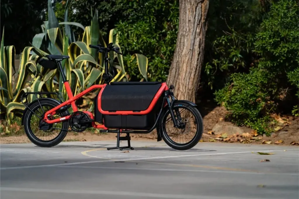 Una bicicleta eléctrica de carga Riese Müller Carrie2 roja está aparcada en una superficie pavimentada cerca de plantas y árboles verdes, mostrando su diseño compacto.