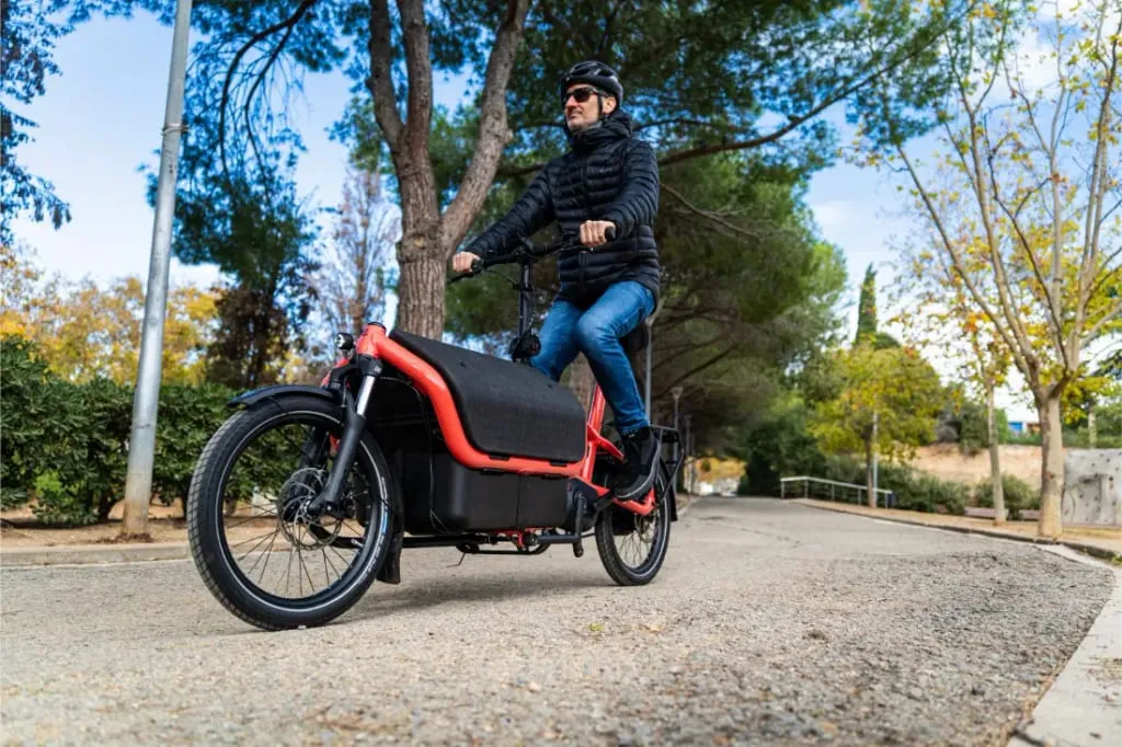 Un hombre con casco monta en una bicicleta de carga Riese Müller roja de tamaño compacto por un sendero arbolado de un parque.