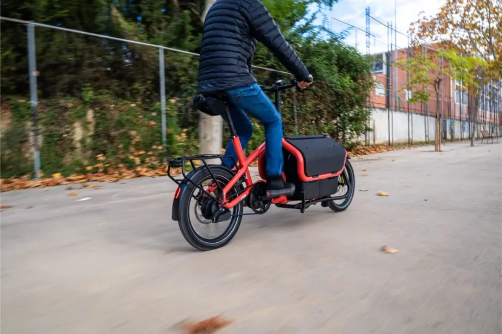 Una persona conduce una Riese Müller Carrie2 roja, una bicicleta eléctrica de carga automática y compacta, por un camino pavimentado flanqueado por árboles y una valla.