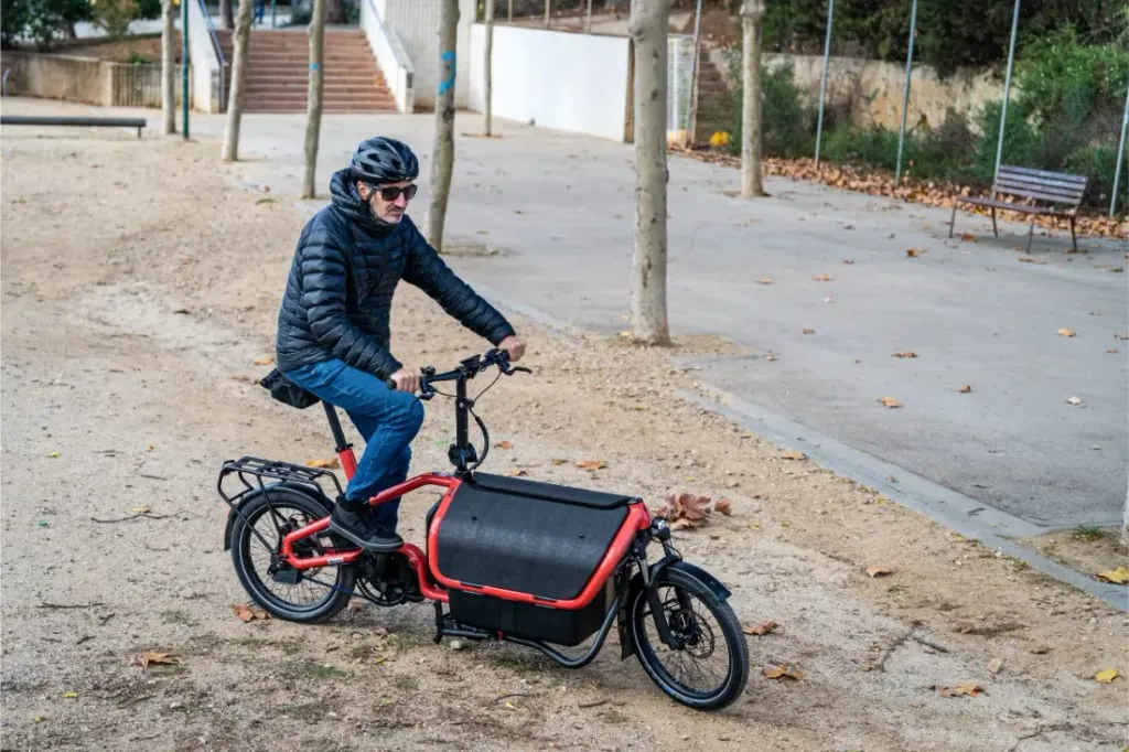 Una persona con casco monta en una bicicleta de carga Riese Müller Carrie2 roja por un camino de arena en un parque con hojas esparcidas, disfrutando de su diseño compacto y su facilidad automática.