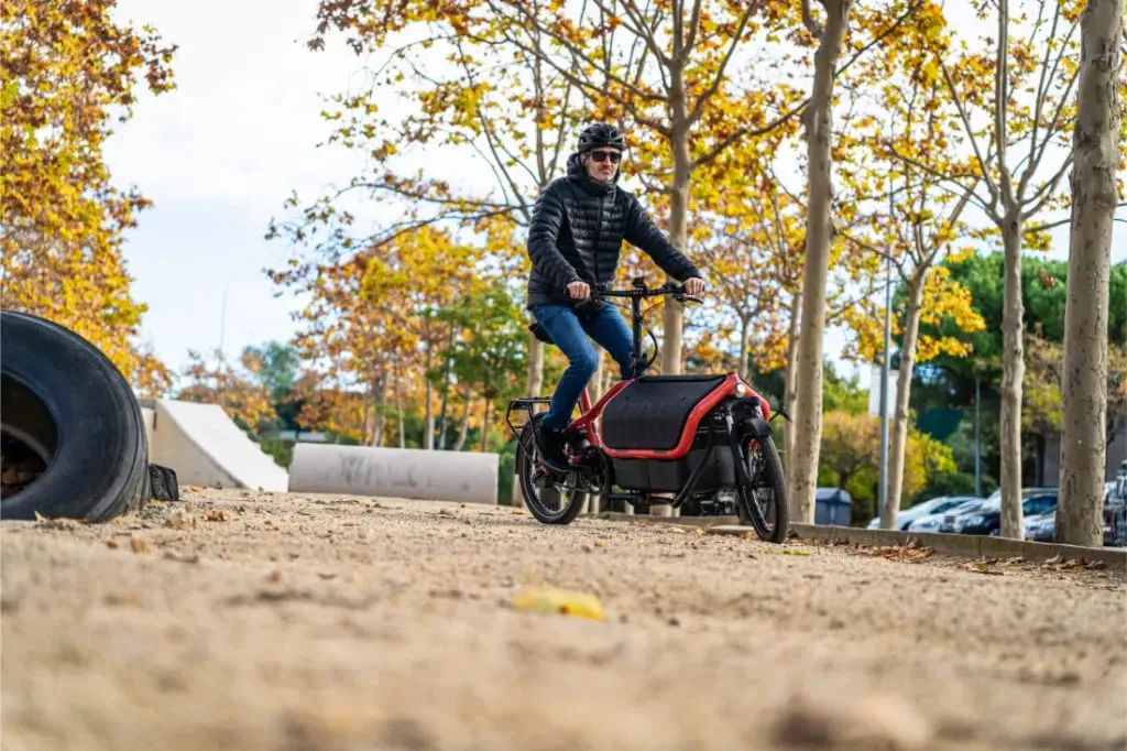 Una persona conduce una bicicleta de carga automática Riese Müller Carrie2 de tamaño compacto por un camino de arena bordeado de árboles otoñales, con casco y chaqueta.