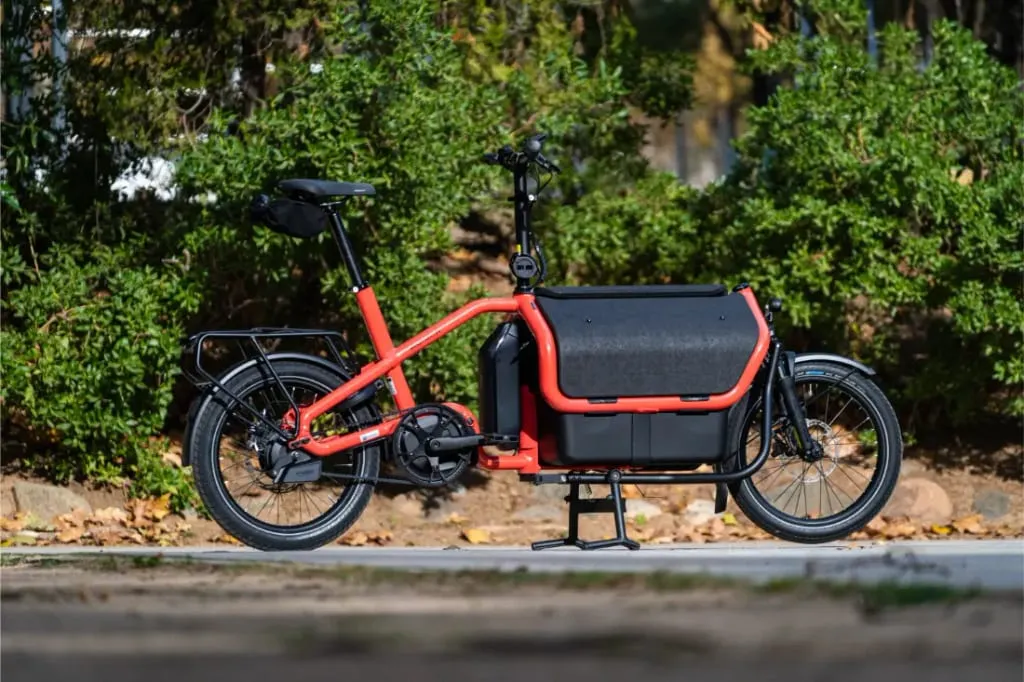 Una bicicleta de carga eléctrica automática Riese Müller Carrie2 roja con ruedas negras está aparcada en un camino pavimentado delante de arbustos verdes, mostrando su tamaño compacto y su diseño moderno.