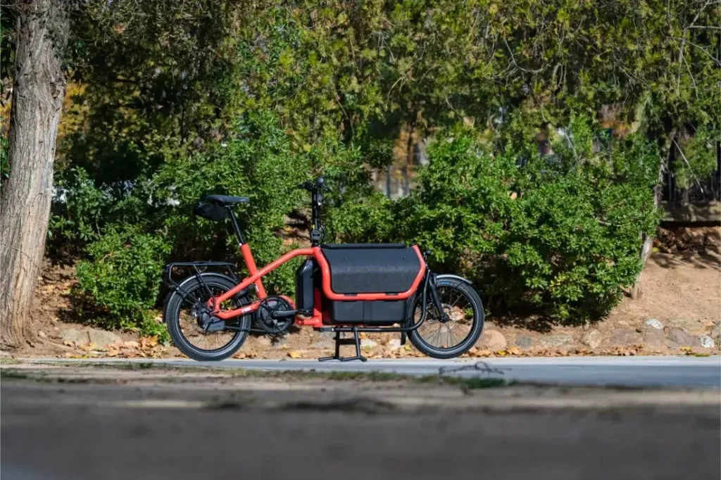 Una bicicleta de carga Riese Müller Carrie2 de color rojo con detalles en negro está aparcada en un camino pavimentado frente a arbustos y árboles verdes. Su diseño compacto la hace perfecta para circular por espacios urbanos.