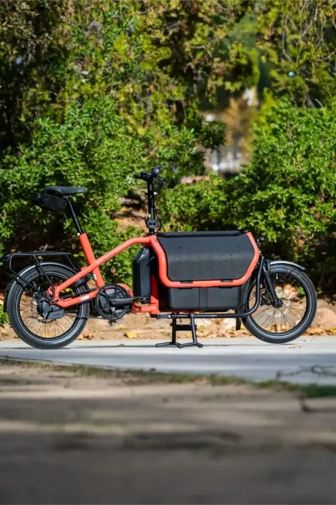 Una bicicleta de carga eléctrica automática Riese Müller Carrie2 de color rojo con tamaño compacto y un gran compartimento de almacenamiento delantero está aparcada en un camino al aire libre.