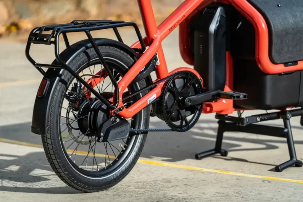 Primer plano de la rueda trasera y la zona de pedales de una bicicleta de carga eléctrica Riese Müller Carrie2 roja aparcada en el pavimento, destacando su diseño compacto.