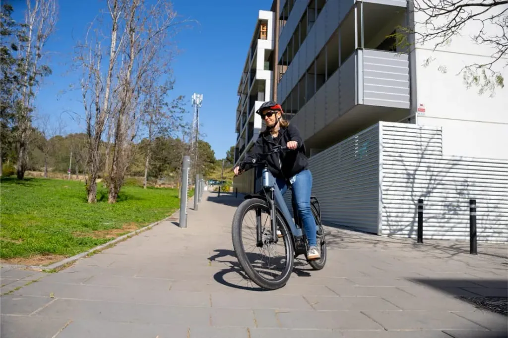Mujer montando en una bicicleta Riese & Müller Nevo5 en una acera cerca de edificios modernos y una zona de parque con césped, disfrutando tanto del lujo accesible como de la silenciosa tecnología CORE en un día soleado.
