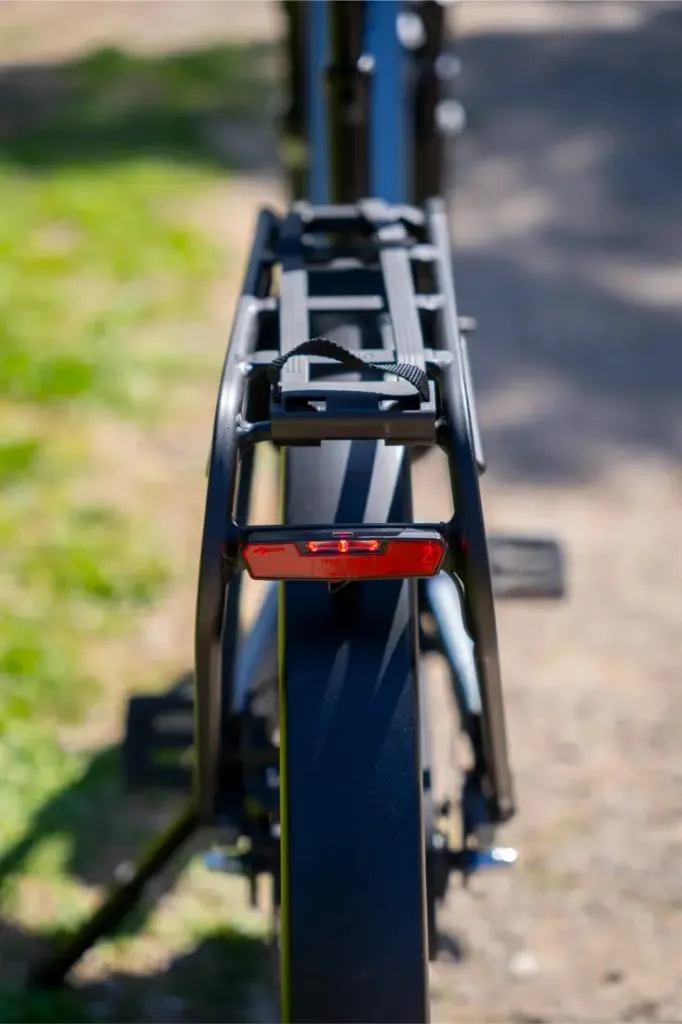 Vista trasera de una bicicleta Nevo5 negra de Riese & Müller con portaequipajes y luz trasera roja, aparcada en un camino iluminado por el sol, donde el lujo accesible se une al elegante diseño urbano.