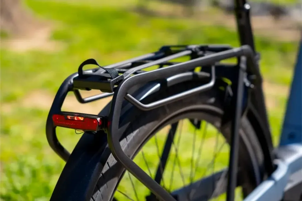Primer plano de la rueda trasera de una Riese & Müller Nevo5 con un portaequipajes negro y un reflector rojo, mostrando el silencioso rendimiento del CORE al aire libre en un día soleado, perfecto para los que buscan un lujo accesible.