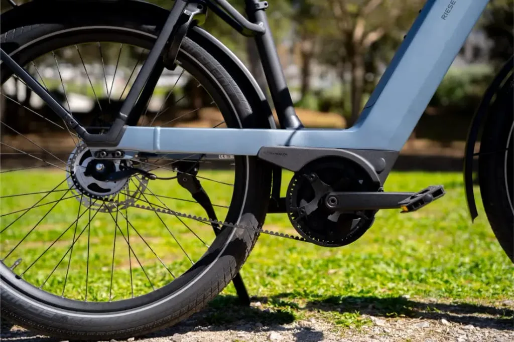 Primer plano de la rueda trasera, la transmisión y los pedales de una bicicleta eléctrica Nevo5 de Riese & Müller, que exhibe su silencioso sistema CORE, aparcada sobre hierba: un toque de lujo accesible para los trayectos cotidianos.