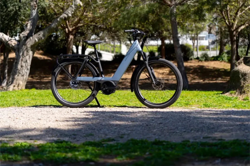 Una bicicleta eléctrica Riese & Müller Nevo5 de color azul claro con tecnología CORE silenciosa aparcada en un camino de grava en un parque con árboles y hierba verde, ofreciendo lujo accesible para cada paseo.