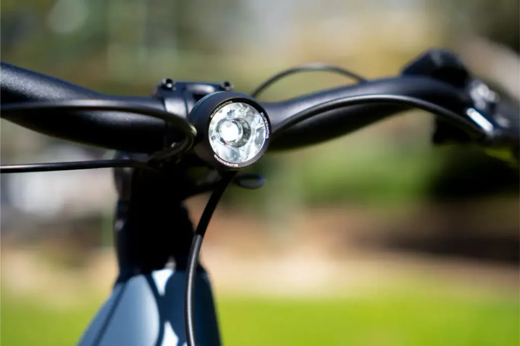 Primer plano de un manillar de bicicleta Riese & Müller Nevo5 con un faro redondo, fondo borroso verde y marrón, que muestra el lujo accesible para los ciclistas urbanos.