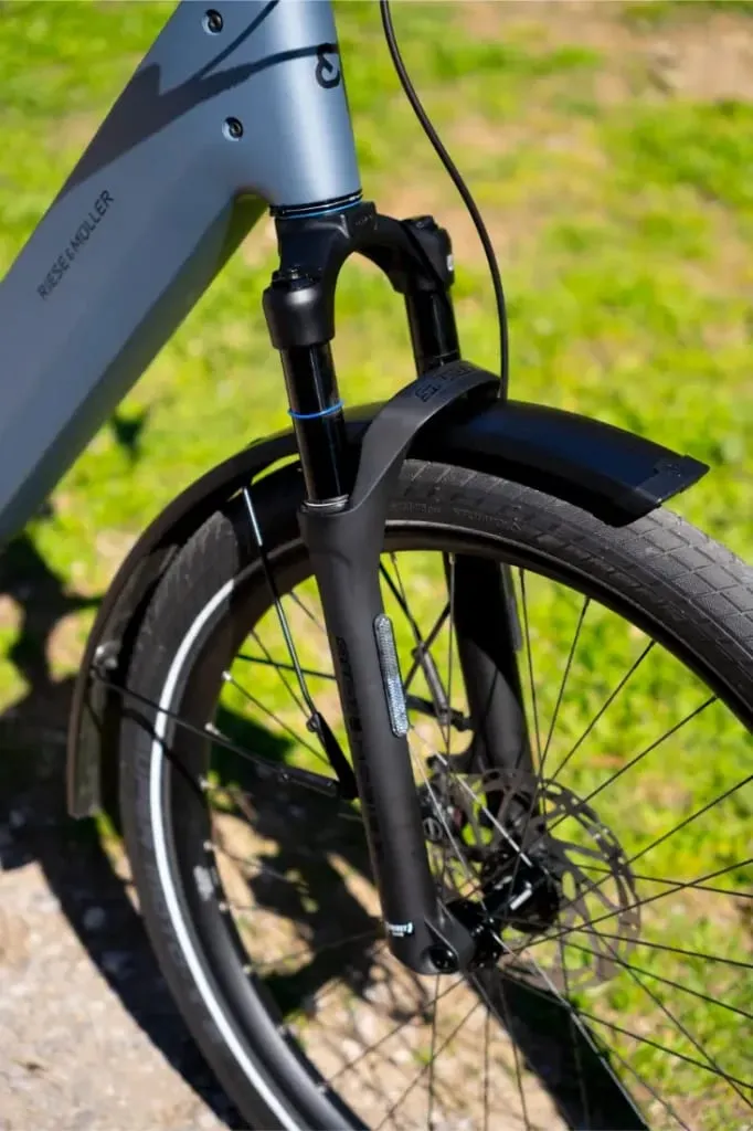 Primer plano de una moderna rueda delantera de bicicleta Riese & Müller Nevo5, horquilla de suspensión y guardabarros sobre un fondo de hierba: un ejemplo de lujo accesible y silencioso rendimiento CORE.