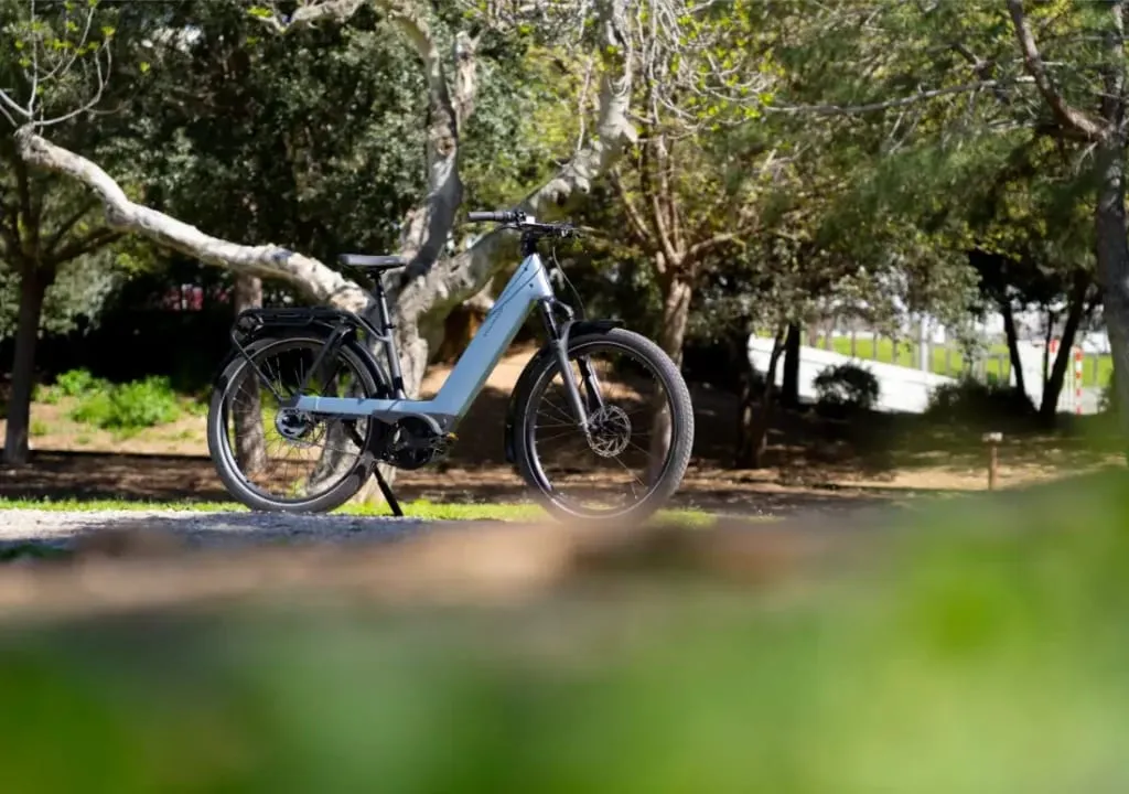 Una bicicleta eléctrica Riese & Müller Nevo5 plateada, con tecnología CORE silenciosa, está aparcada en un sendero de un parque soleado y arbolado.