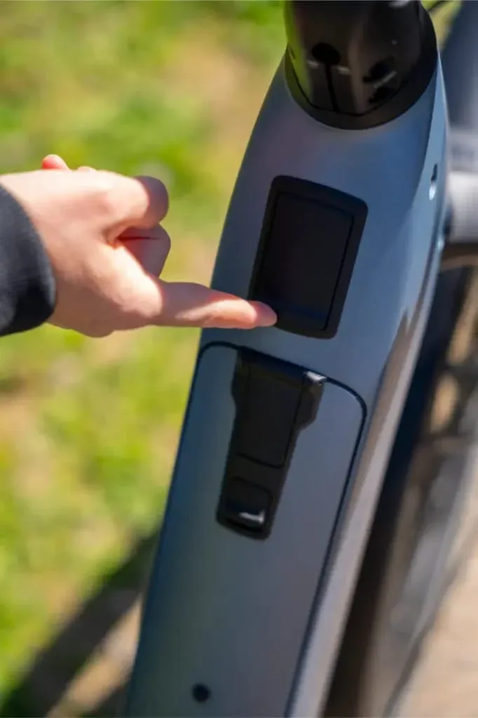 Una persona pulsa un botón o una pantalla en el cuadro de una bicicleta eléctrica Nevo5 gris de Riese & Müller al aire libre, experimentando el lujo accesible y la suave conducción de la silenciosa tecnología CORE.
