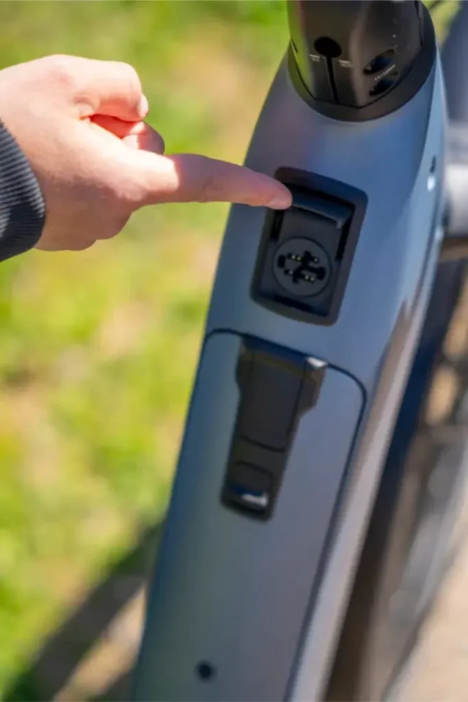 Una mano señala el puerto de carga en el cuadro de una bicicleta eléctrica Nevo5 de Riese & Müller al aire libre, destacando el lujo accesible y la silenciosa tecnología CORE.