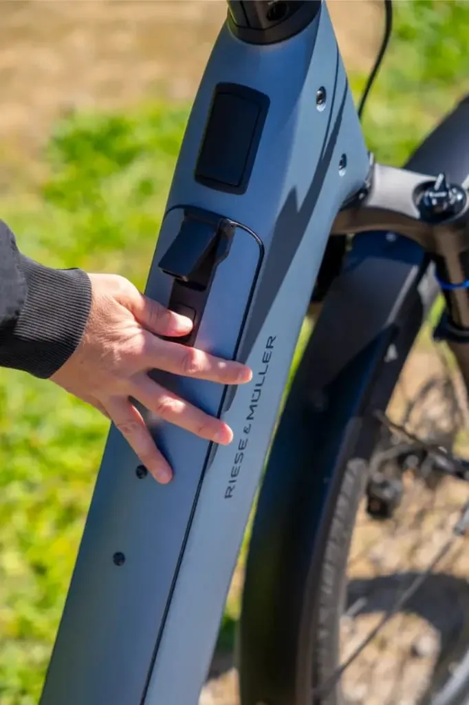 Una mano toca el compartimento de la batería de una bicicleta eléctrica Nevo5 de Riese & Müller al aire libre, destacando la silenciosa tecnología CORE para una experiencia accesible de lujo.
