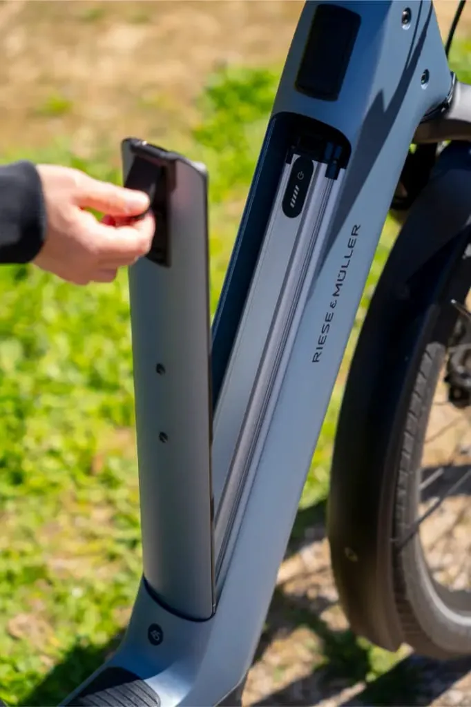 Hand removing battery from a Riese & Müller Nevo5 bicicleta eléctrica frame, with green grass in the background-una muestra de lujo accesible en cada detalle.