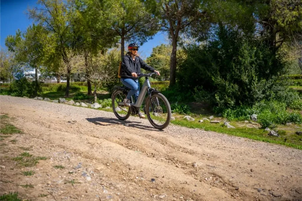 Una persona conduce una Riese & Müller Nevo5 por un camino de tierra rodeado de árboles y vegetación, disfrutando del lujo accesible y del silencioso CORE en un día soleado.