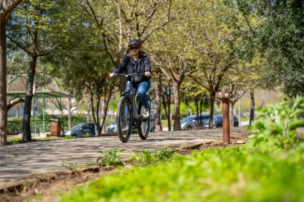 Una persona monta en una bicicleta Nevo5 de Riese & Müller por un camino soleado y arbolado, con hierba verde y coches aparcados al fondo, disfrutando del lujo accesible en cada momento de silencio CORE.