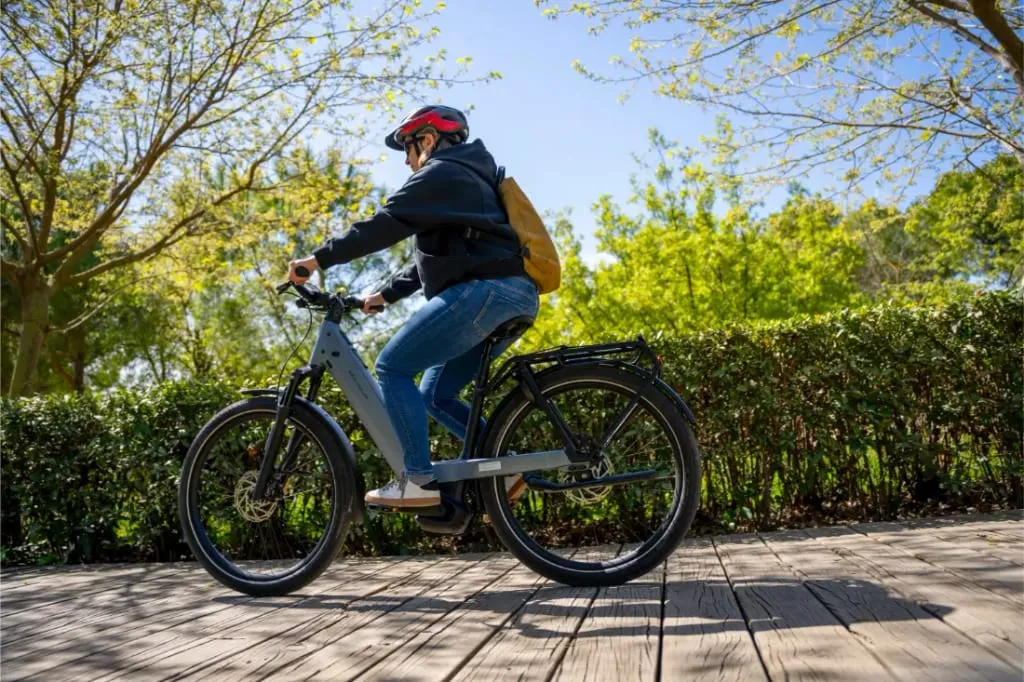 Persona con casco y mochila monta en una bicicleta Riese & Müller Nevo5 con tecnología CORE silenciosa por un sendero de madera rodeado de vegetación y árboles, disfrutando del lujo accesible en la naturaleza.