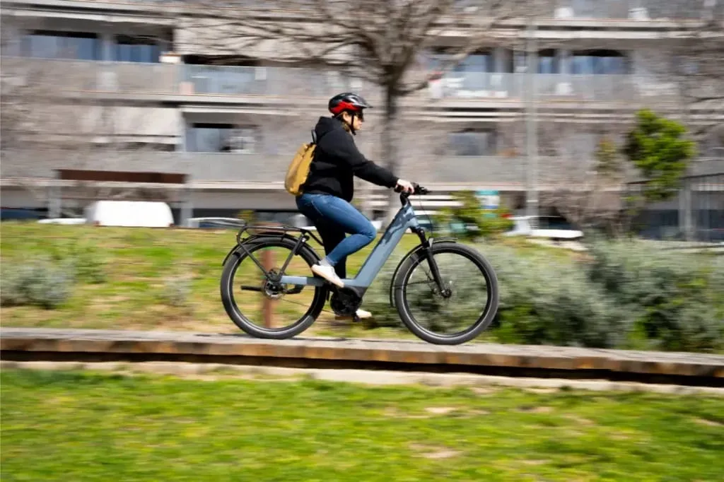 Persona con casco conduce una bicicleta eléctrica Riese & Müller Nevo5 por un parque, disfrutando del lujo accesible mientras los edificios se difuminan en el fondo.
