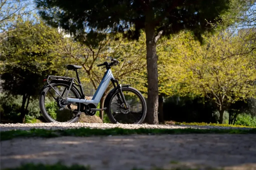Una bicicleta eléctrica Riese & Müller plateada se encuentra en un camino de grava bajo un árbol en un parque iluminado por el sol, mostrando el lujo accesible y la tecnología CORE silenciosa Nevo5.