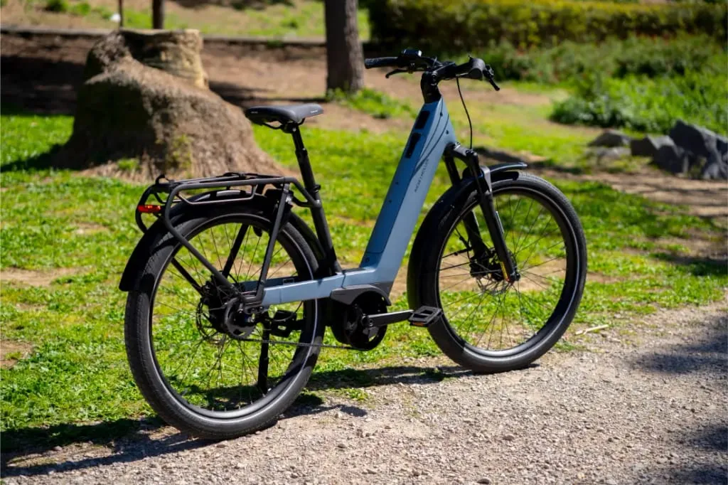 Una bicicleta eléctrica Riese & Müller Nevo5 de color azul claro con tecnología CORE silenciosa aparcada en un camino de grava en un parque soleado y verde: perfecta para los que buscan lujo accesible.