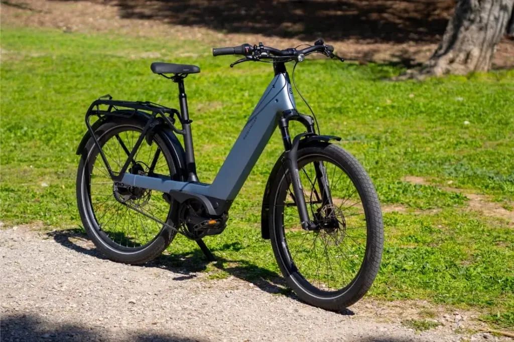 Una moderna bicicleta eléctrica Riese & Müller Nevo5 gris está aparcada en un camino de grava junto a una zona de césped con árboles al fondo, ofreciendo tecnología CORE silenciosa y lujo accesible para cada paseo.