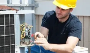 an electrician wearing a yellow hard hat repairing the electrical wiring on an outdoor HVAC system