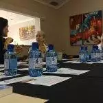 Three women are seated at a table with water bottles, documents, and mugs, engaged in conversation in a room with colorful artwork.