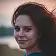 Close-up headshot of a young woman with reddish-brown, curly hair smiling slightly, with a soft outdoor background at sunset.
