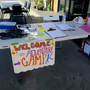 Colorful "Welcome to Adventure Camp!" sign on an outdoor registration table with supplies.
