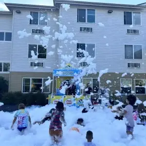 Children gleefully play in a large, bubbly pile of white foam outdoors.